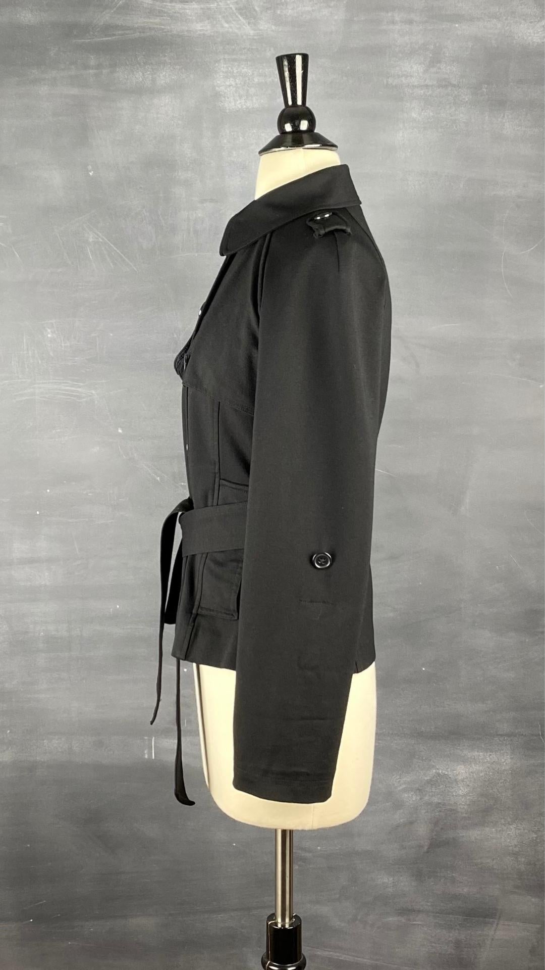 Veste courte noire façon trench. Extensible et polyvalente, les manches se roulent et sont maintenues en place à l'aide d'une sangle. Ceinturée à la taille. Vue de côté, la manche déroulée.