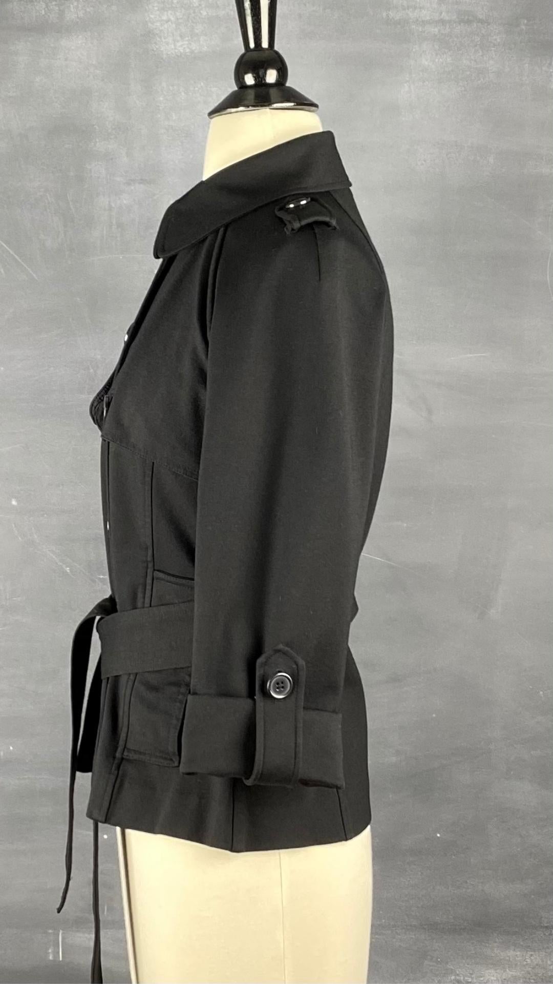 Veste courte noire façon trench. Extensible et polyvalente, les manches se roulent et sont maintenues en place à l'aide d'une sangle. Ceinturée à la taille. Vue de côté, la manche roulée.