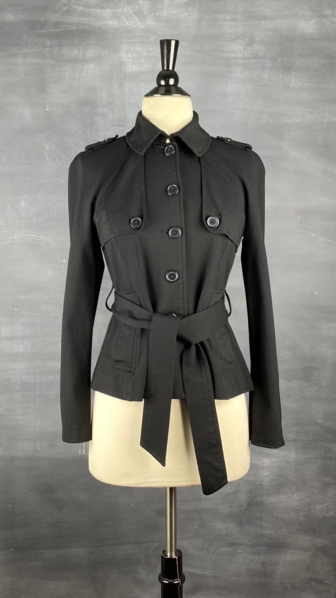 Veste courte noire façon trench. Extensible et polyvalente, les manches se roulent et sont maintenues en place à l'aide d'une sangle. Ceinturée à la taille. Vue de face.