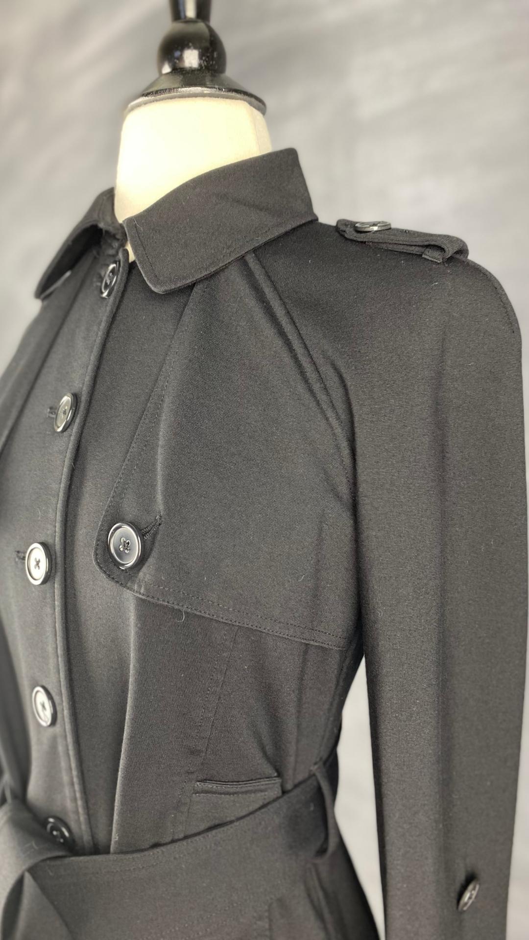 Veste courte noire façon trench. Extensible et polyvalente, les manches se roulent et sont maintenues en place à l'aide d'une sangle. Ceinturée à la taille. Vue légèrement de côté du haut de la veste et des détails.