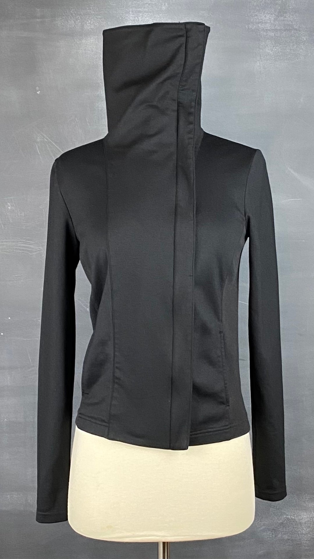Veste noire à grand col zippé, légèrement décentré. Vue de face, col zippé en entier.