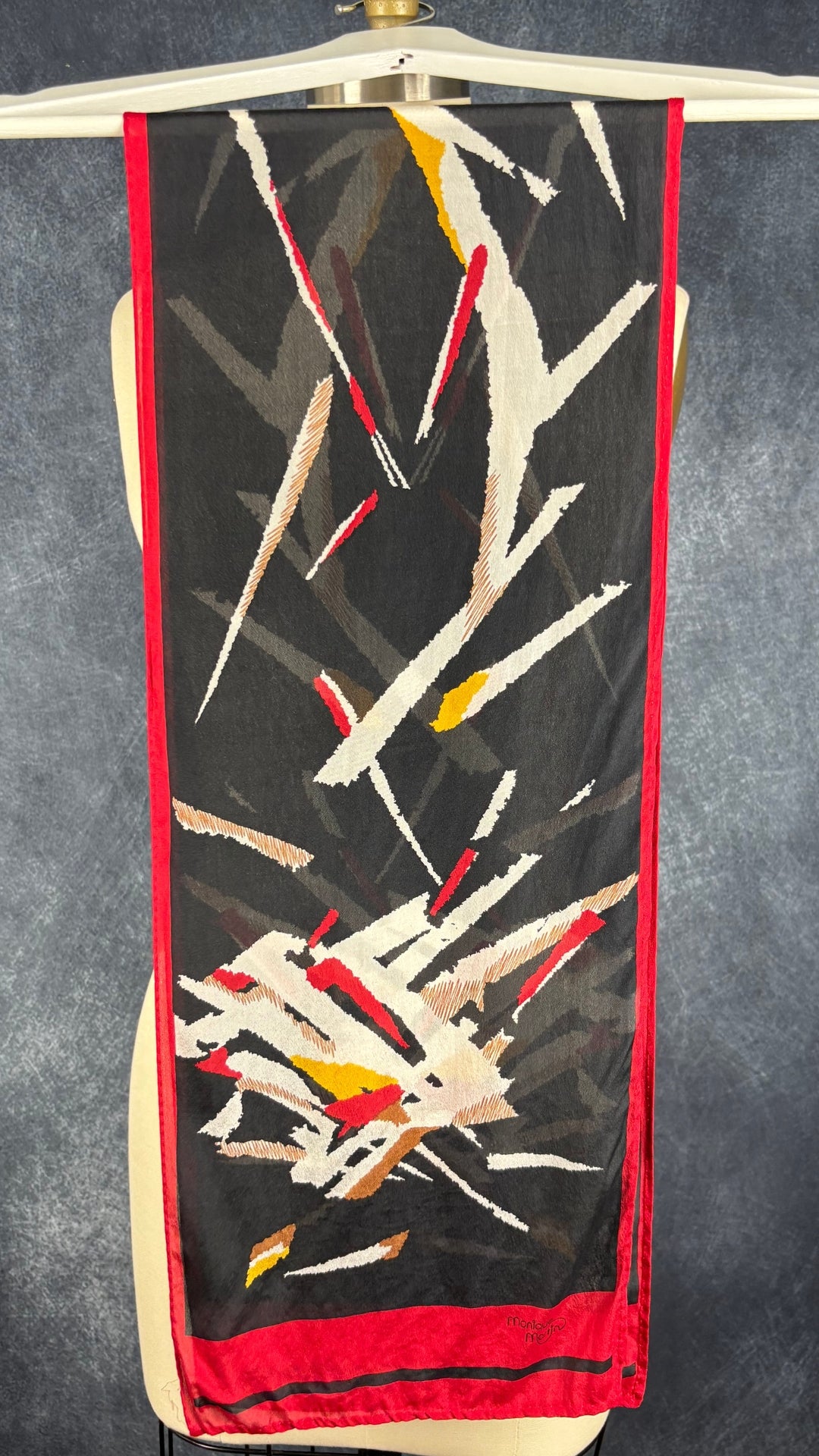 Foulard noir, blanc et rouge Monique Martin, rectangulaire long. Vue du foulard plié en deux sur un cintre, du côté où la marque est inscrite.