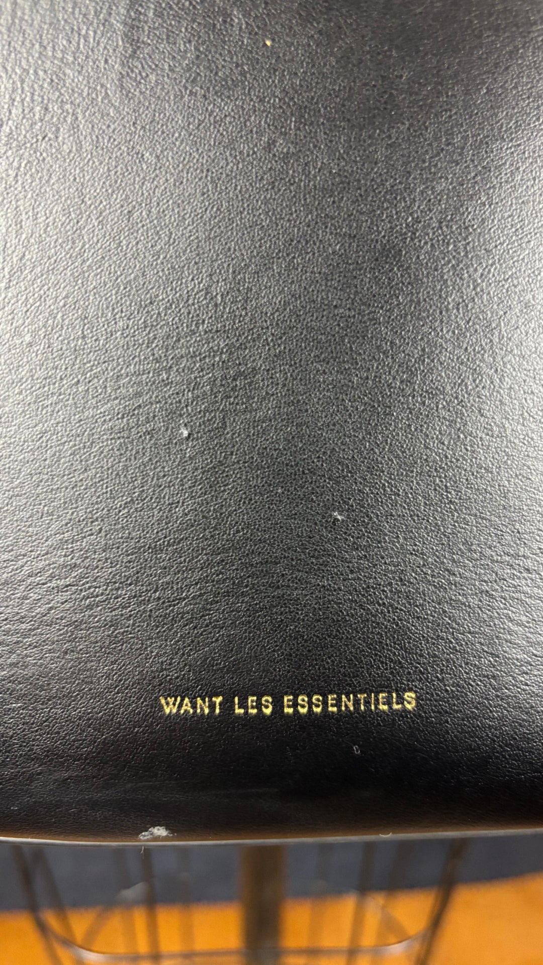 Sac sceau noir à poignée et bandoulière en cuir Want Les Essentiels. Vue de près de la marque gravée en doré sur le devant du sac. Il y a également une petite marque où le cuir est un peu abimé dans le bas du sac.